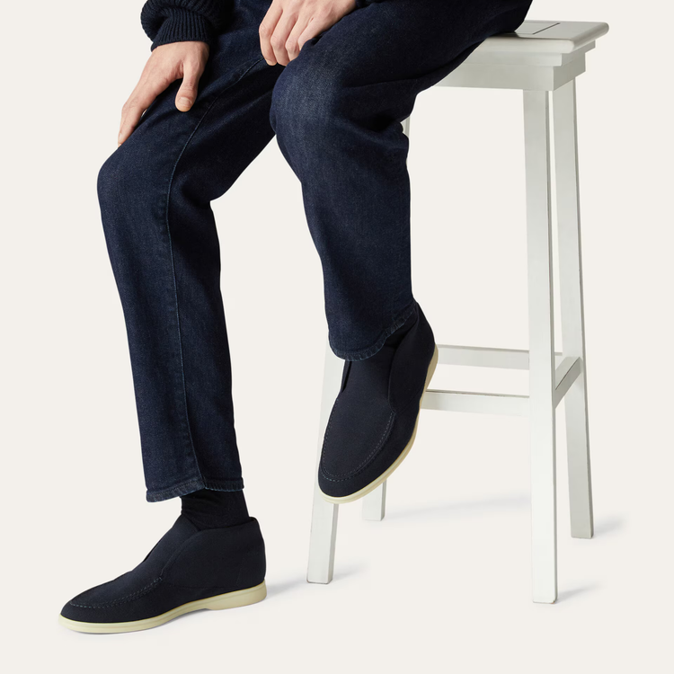 Person wearing navy blue shoes and jeans sitting on a white stool against a white background